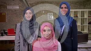 Three smiling arabian women ar standing near camera and looking cwith confidence, wearing different hijab, in the brick