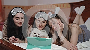 Three smiling young women, with sleep masks on their heads, and a white Spitz, lie on the bed and look at the tablet