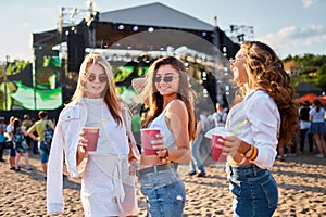 Three smiling young women toast with cups at sunny seaside concert. Group of happy females enjoy summer music event on