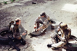 Three soldiers are sitting on tires and having rest. They had a good fight. Men want to keep their strenght for the next