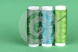 Three spools of sewing thread of different shades of green with a needle stuck in and isolated on green