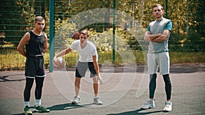 Three sportsmens standing on the basketball court outdoors and looking in the camera - one of them playing with a ball