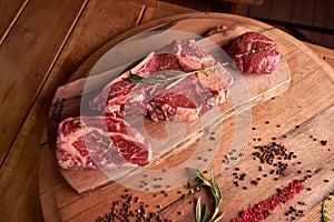 Three steaks of rib eye on a wooden cutting board with a sprig of rosemary, colored peppercorns, dried pickles and a glass of wine