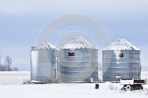 Three steel silos or grain bins covered in snow on a farm in a rural area in winter with a small wagon in the foreground