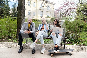 Three students on a bench in the park study talking typing on laptop notebook and writing. Education concept. Young colleagues