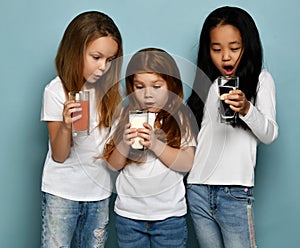 Three surprised socked kids girls friends in white t-shirts stand looking at glasses of water, milk and fresh juice