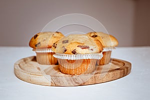 Three tasty muffins with chocolate peices on a wooden tray on a white table