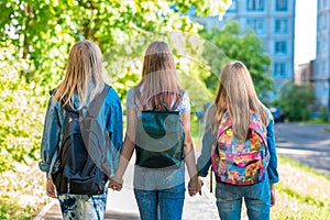 Three teenage girls. They hold each other`s hands. Returns home after school. Summer in nature. Behind backpacks. Back