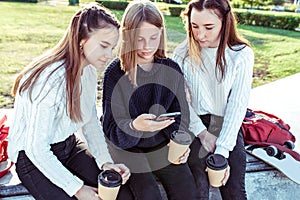 Three teenage girls schoolgirls 12-14 years old, in summer in city, in hand smartphone, cup tea drink, watch photos on