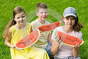 Three teenagers eat slices of watermelon sitting on green grass