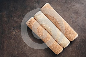 Three terry beige towels rolled up on a dark brown rustic background. Top view, flat lay, copy space