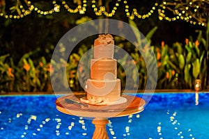 Three tier layered wedding cake with white marzipan icing decorated with orchids on a table near the pool.