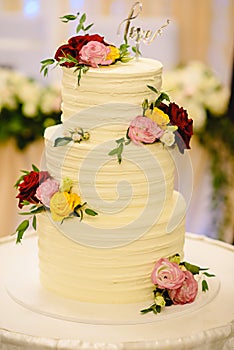 Three-tiered white wedding cake decorated with flowers from mastic on a white wooden table.