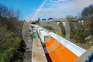 Three trains riding on the railroad tracks surrounded by lush green trees and bare winter trees and clouds, blue sky and a rainbow