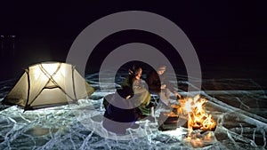 Three travelers by fire right on ice at night. Campground on ice. Tent stands next to fire. Lake Baikal. Nearby there is