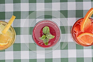Three tropical mixed drinks, berries, orange and kiwi fruit cocktails served on green plaid tablecloth.