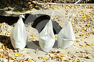 Three white plastic bags full of sand in front of the trench on the street reconstruction site and with orange autumn leaves aroun