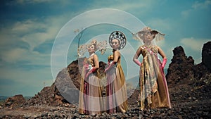 Three women in artistic costumes with headpieces standing in a rocky landscape, portraying a fantasy or tribal scene