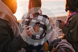 Three women female friends having picnic, drinking hot tea from iron mugs, sitting on beach