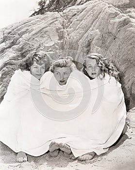Three women huddled under a towel on beach
