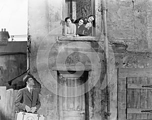 Three women looking out of a window at a man standing in the street with a suitcase