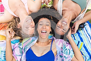 Three women are lying on a colorful blanket, heads together, smiling joyfully