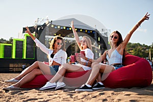 Three women relax on red bean bag chairs at beach music festival, sunny day, raising drinks, stage in background