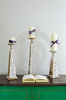 Three wooden candlesticks with engraved inscriptions holding three candles and opened book on green vintage wooden sideboard