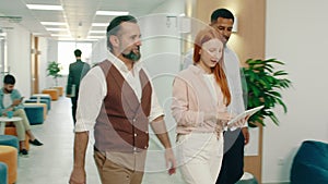 Three work colleagues are walking together to a table in the office, they greet people as they walk by, when they sit at