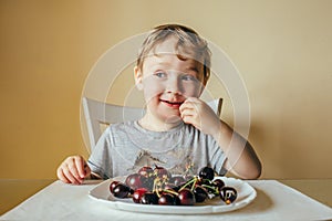 A three-year-old child sits in the kitchen of the house and eats cherries. he had a sly look aside.