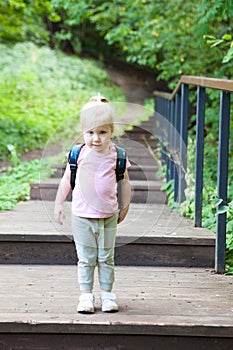 Three-year-old girl with a backpack stands on a wooden ladder in the park