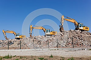 Three yellow excavators on a pile of dirt on behind the fence