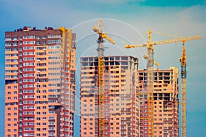 Three yellow tower cranes and apartment buildings under construction against the blue sky. Construction of multi-storey buildings