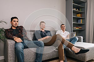 Three young adult friends are sitting on soft couch and watching TV