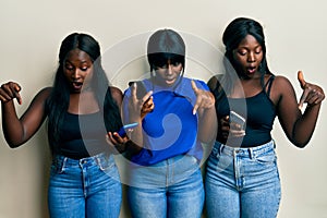 Three young african american friends using smartphone pointing down with fingers showing advertisement, surprised face and open