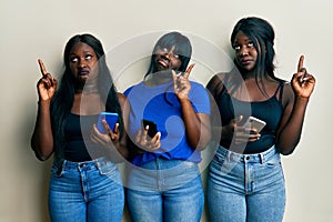 Three young african american friends using smartphone pointing up looking sad and upset, indicating direction with fingers,