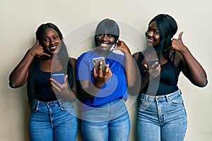 Three young african american friends using smartphone smiling doing phone gesture with hand and fingers like talking on the