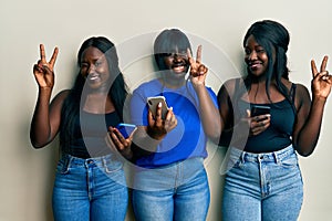 Three young african american friends using smartphone smiling with happy face winking at the camera doing victory sign with