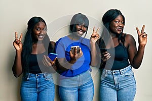 Three young african american friends using smartphone smiling looking to the camera showing fingers doing victory sign