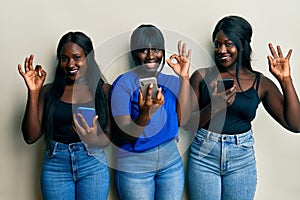 Three young african american friends using smartphone smiling positive doing ok sign with hand and fingers