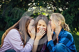 Three young attractive woman sharing secrets sitting on green grass in the park. Cheerful girlfriends gossip and whisper outdoor