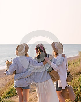 Three young beautiful women female friends going to have summer picnic on a beach at sunset