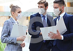 Three young businessmen wear preventive masks during epidemy, stand and discuss business at an office meeting.