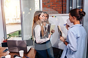 Three young businesswomen sharing new business ideas holding notes standing at whiteboard in office