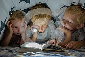 Three young children are reading a book with a flashlight under the covers at night.