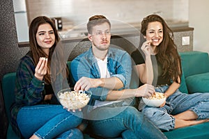 Three young friends two beautiful women and one handsome man watching tv on the sofa eating pop corn, friendship