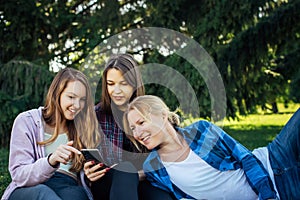 Three young girls sitting on green grass with book and smartphones. Cheerful friends resting in park. Spring and summer leisure