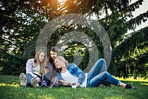 Three young girls sitting on green grass with book and smartphones. Cheerful friends resting in park. Spring and summer leisure