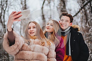 Three young happy friends, two girls and boy making selfie on mobile phone against winter landscape