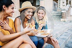 Three young italian women eating pizza sitting together outdoors.